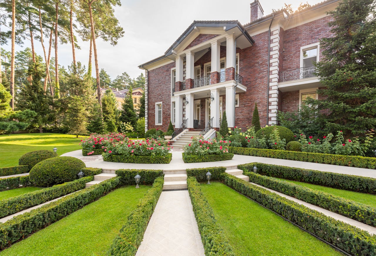 Luxurious brick mansion surrounded by lush gardens and trimmed hedges on a sunny day.
